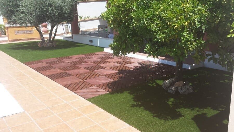 Patio soleado al aire libre con pavimento estampado en rojo, césped verde y árboles junto a un camino pavimentado con baldosas