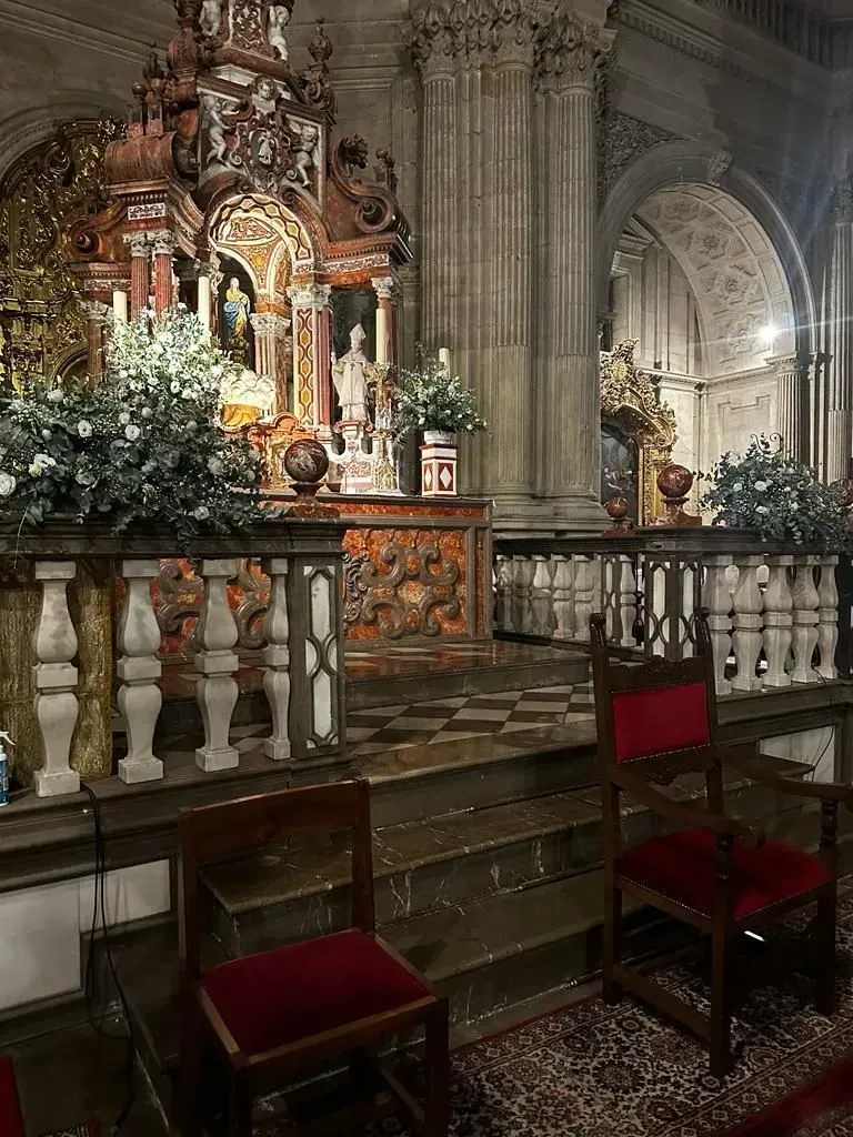 Altar de iglesia ornamentado, con columnas de mármol, flores, velas y sillas con cojines rojos en primer plano