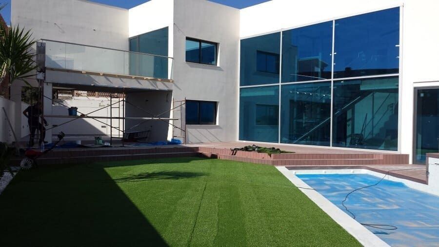 Patio de una casa moderna con césped verde, piscina de azulejos azules y grandes ventanales de cristal reflectante