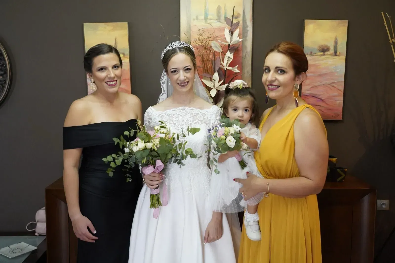 Tres mujeres en una boda, una de ellas con un vestido blanco y un ramo de flores, con un niño delante de ellas.