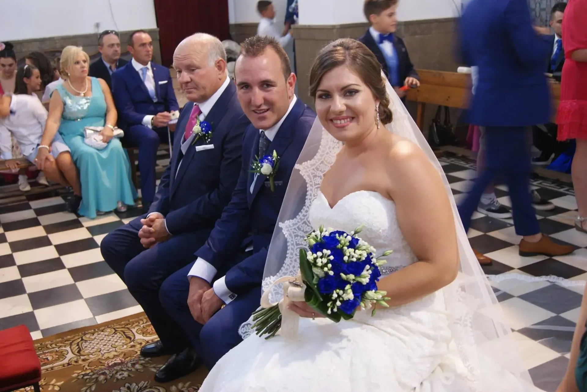 La novia y el novio sentados con un hombre mayor durante una ceremonia de boda, sonriendo en el pasillo de una iglesia