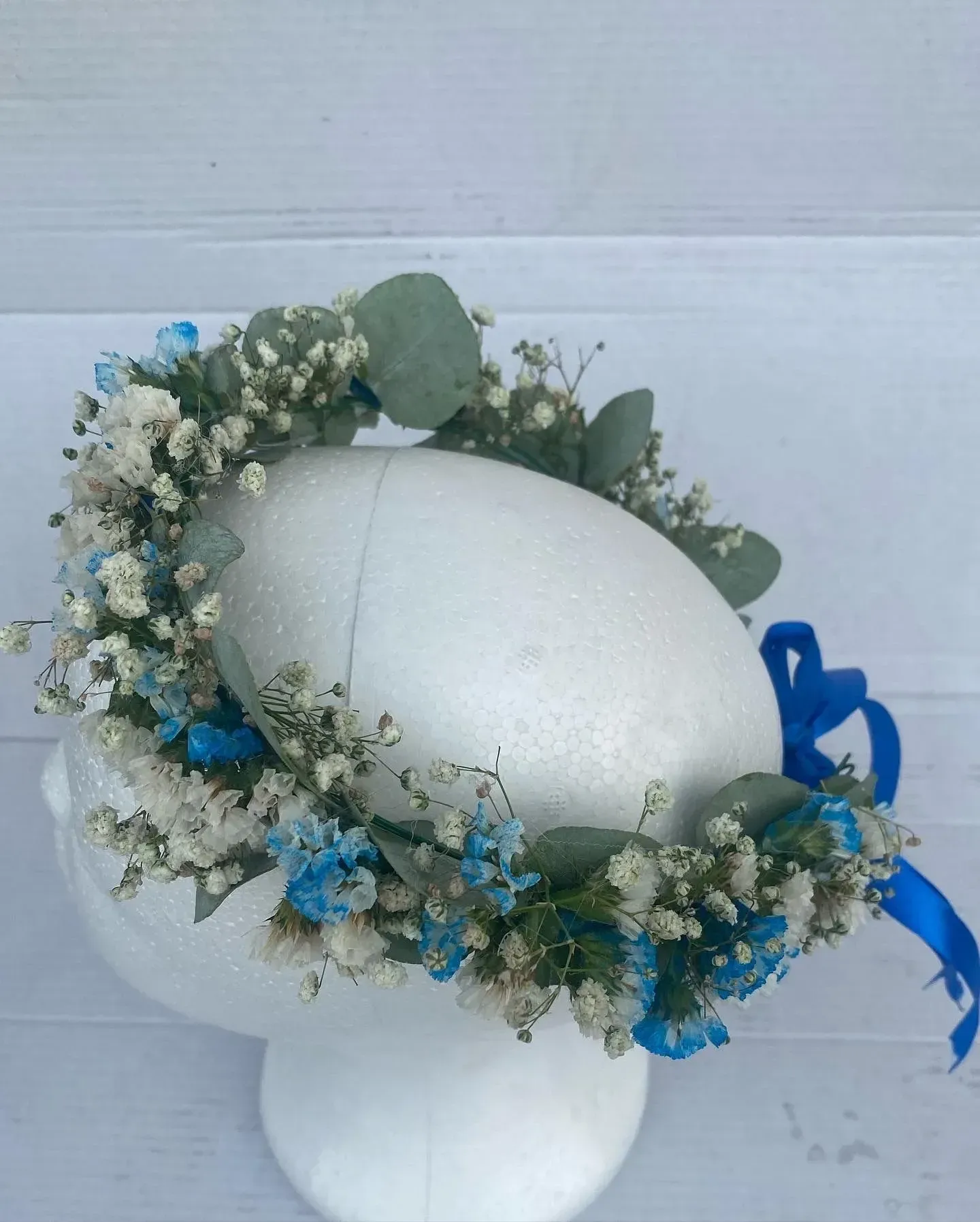 Cabeza de maniquí de espuma blanca con una corona floral azul y blanca y una cinta azul sobre un fondo blanco