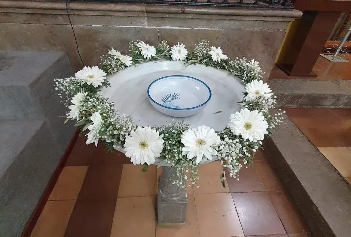 Corona de flores blancas con un cuenco en el centro sobre un soporte de azulejos, en un interior parecido a una iglesia