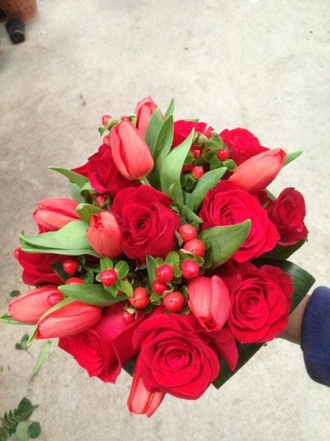 Ramo de rosas rojas y tulipanes rosas con hojas verdes, sostenido en una mano sobre un suelo claro.