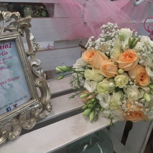 Arreglo floral con rosas color melocotón y blancas junto a un marco plateado ornamentado sobre una mesa de exposición