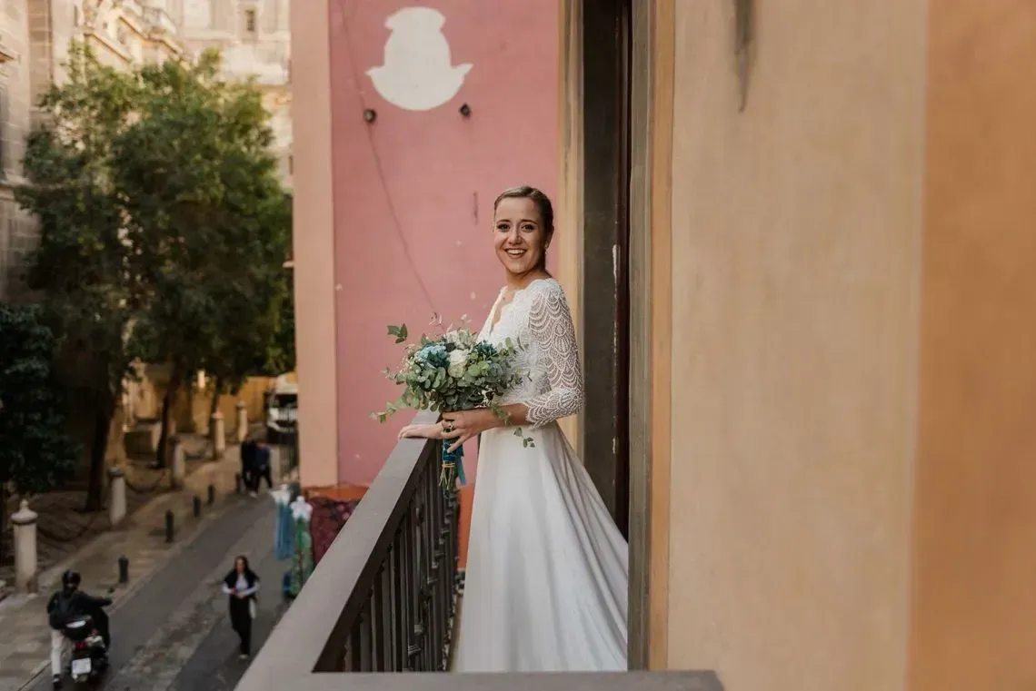 Novia con un vestido blanco y un ramo en la mano, en un balcón con una pared rosa y la calle abajo