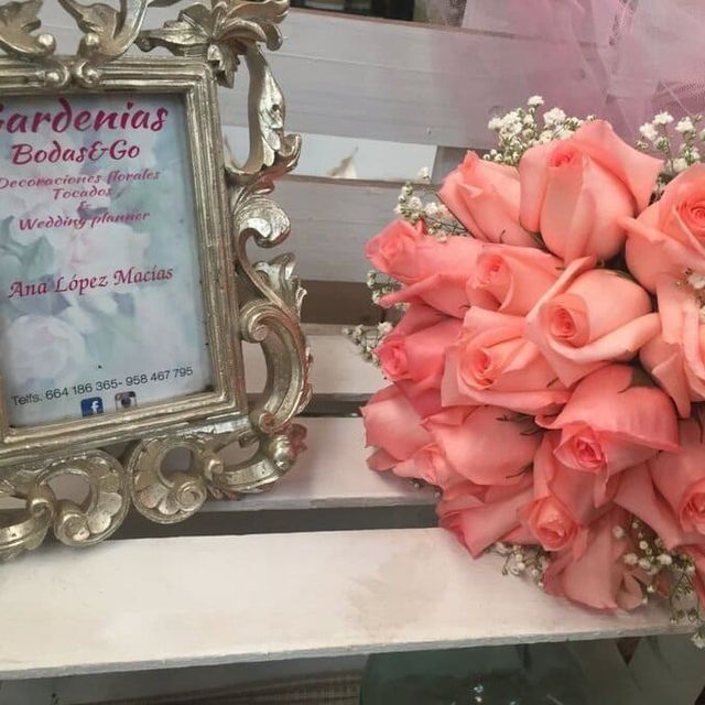 Ramo de rosas rosas junto a un marco plateado ornamentado sobre una estantería blanca, con un letrero de Gardenias Bodas y Co.