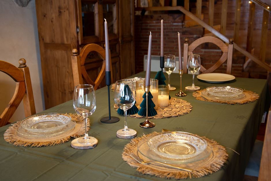 Mesa de comedor preparada para comer con velas, cubiertos y copas de vino sobre un mantel verde en una habitación de madera.