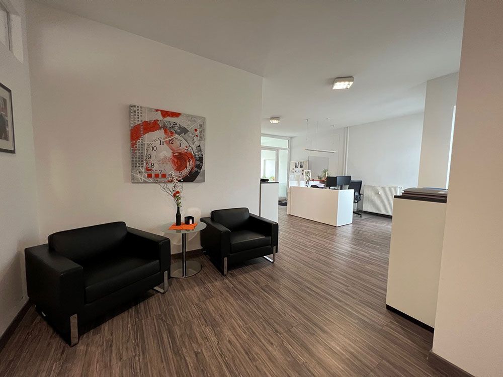 Wartebereich im Büro mit zwei schwarzen Sesseln, Kunstwerken und Boden in Holzoptik. Weiße Wände und ein entfernter Büroraum sind sichtbar.
