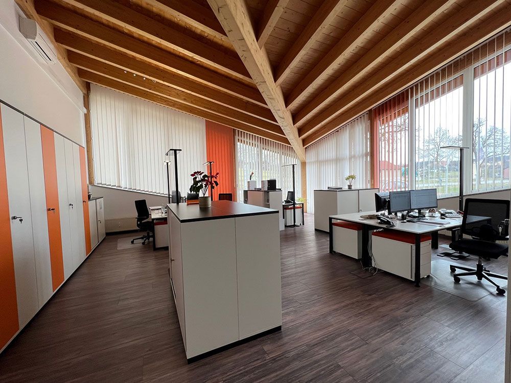 Büroeinrichtung mit Holzbalkendecke, Schreibtischen, Stühlen und Schränken. Weiß-, Orange- und dunkle Holztöne.