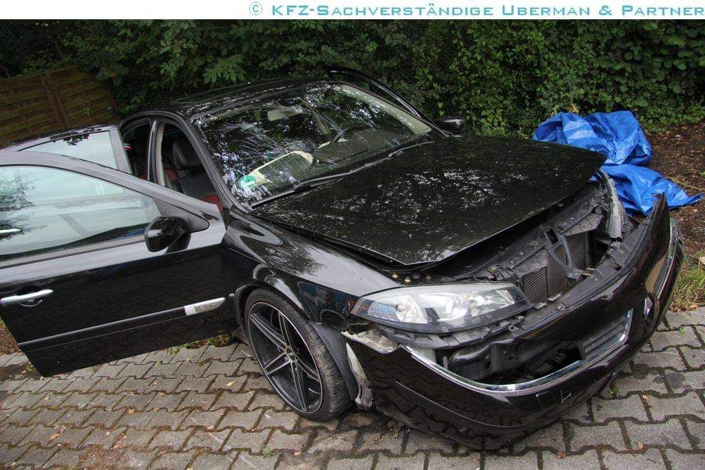 Ein schwarzes Auto mit offener Motorhaube steht nach einem Autounfall auf einer gepflasterten Einfahrt.