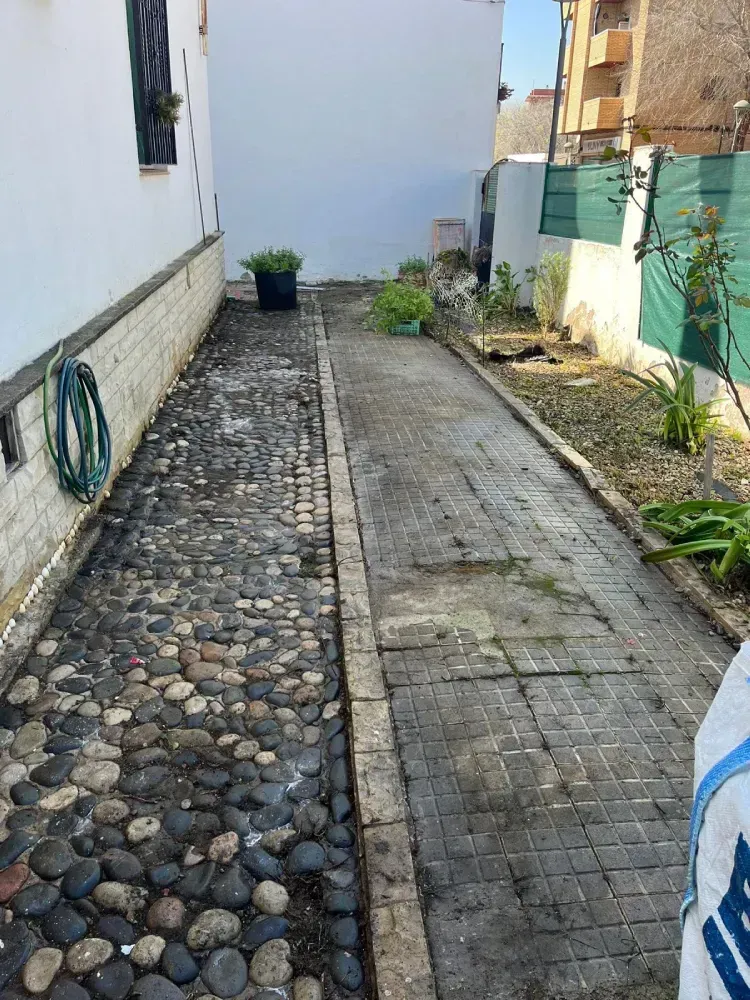 Un estrecho sendero exterior situado entre la pared blanca de un edificio y una valla verde.