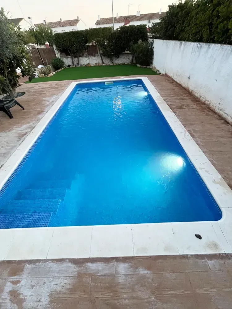 Una piscina rectangular en el patio trasero, con agua azul e iluminación subacuática, rodeada de pavimento de piedra.