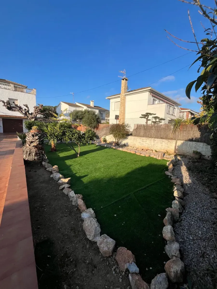 Un patio trasero soleado con un trozo de césped verde bordeado de rocas, rodeado por un camino de grava y casas blancas.
