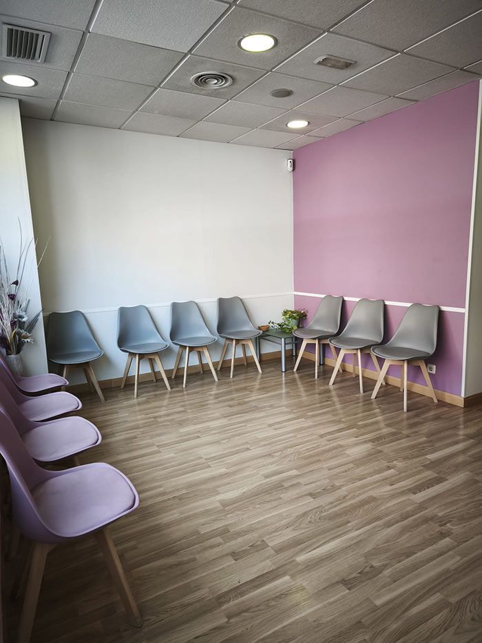 Sala de espera vacía con sillas de color lavanda y gris sobre paredes blancas y rosas, suelo de imitación madera.