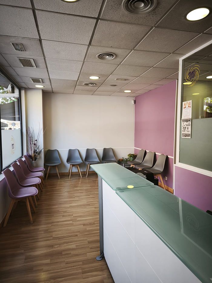 Sala de espera vacía con sillas, una pared decorativa de color rosa y un mostrador de recepción bajo luces de techo.