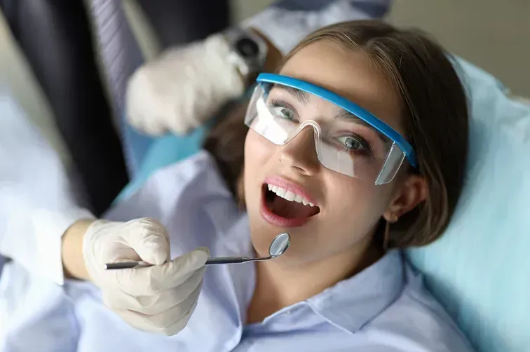 Paciente sonriente con gafas de seguridad durante un examen dental mientras el dentista sostiene un espejo.