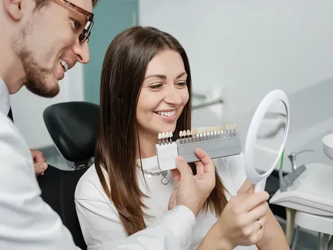 Dentista y paciente examinando muestras de color de dientes en una clínica, con un espejo cerca.
