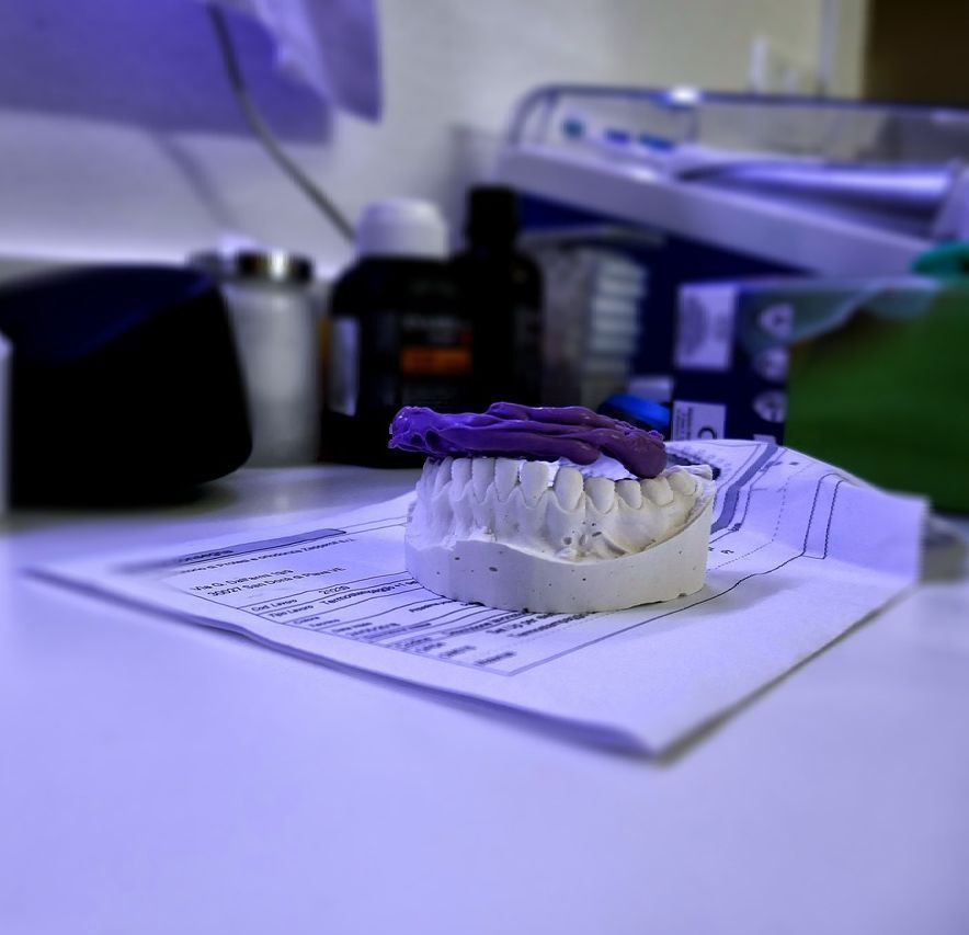 Pequeño objeto impreso en 3D de color morado y blanco que descansa sobre papeles en un escritorio, con material de oficina borroso al fondo.