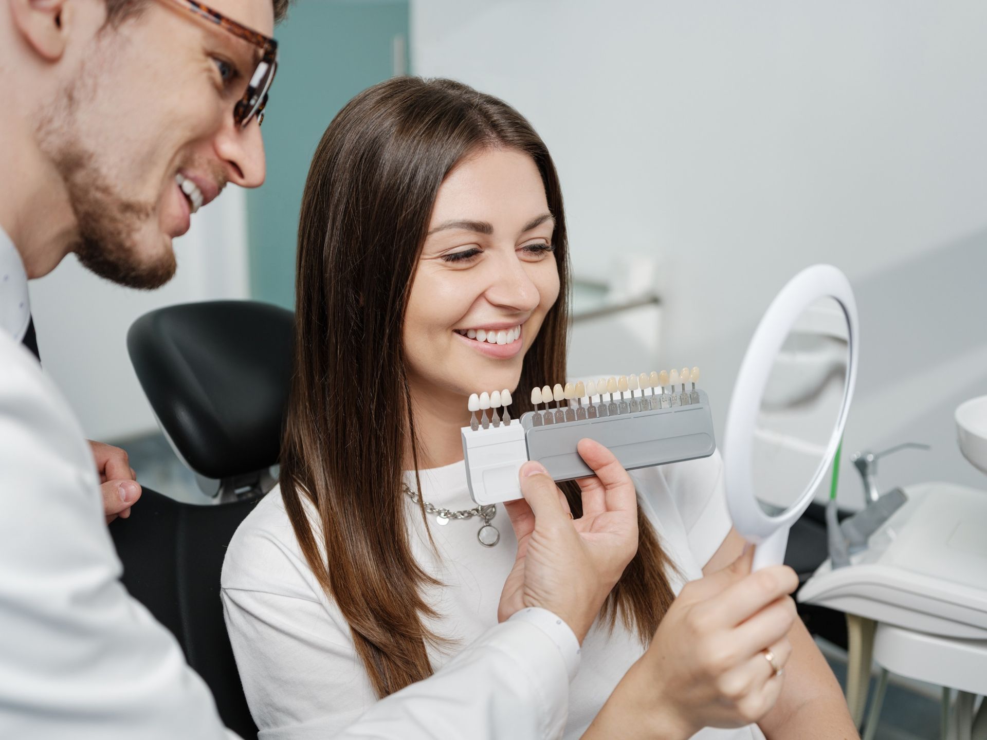 El dentista y el paciente repasan juntos la guía de colores dentales, sonriendo en una clínica luminosa con espejo.