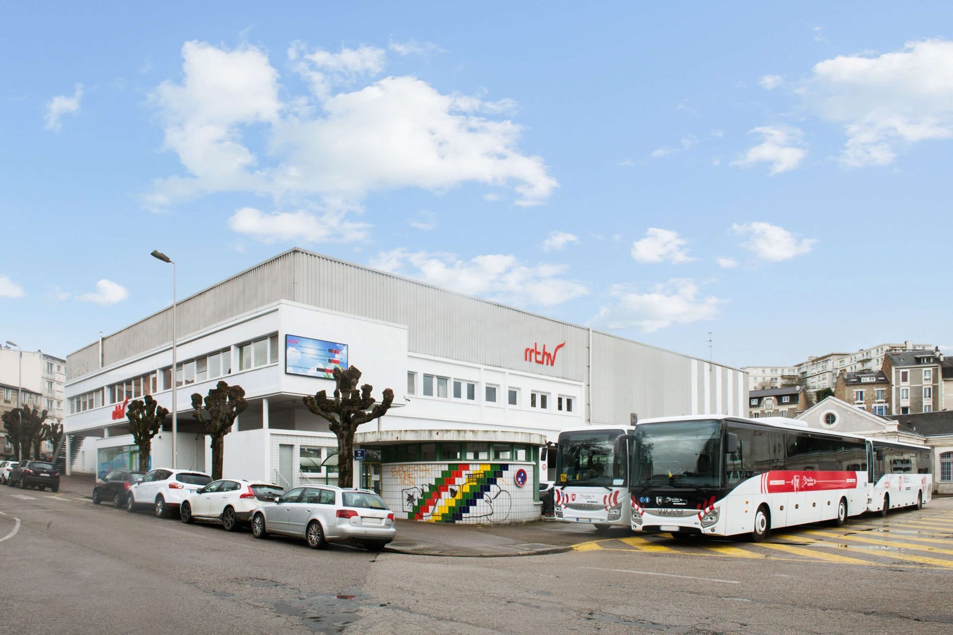 Des bus stationnés devant un bâtiment industriel blanc orné d'un logo rouge, par temps nuageux.