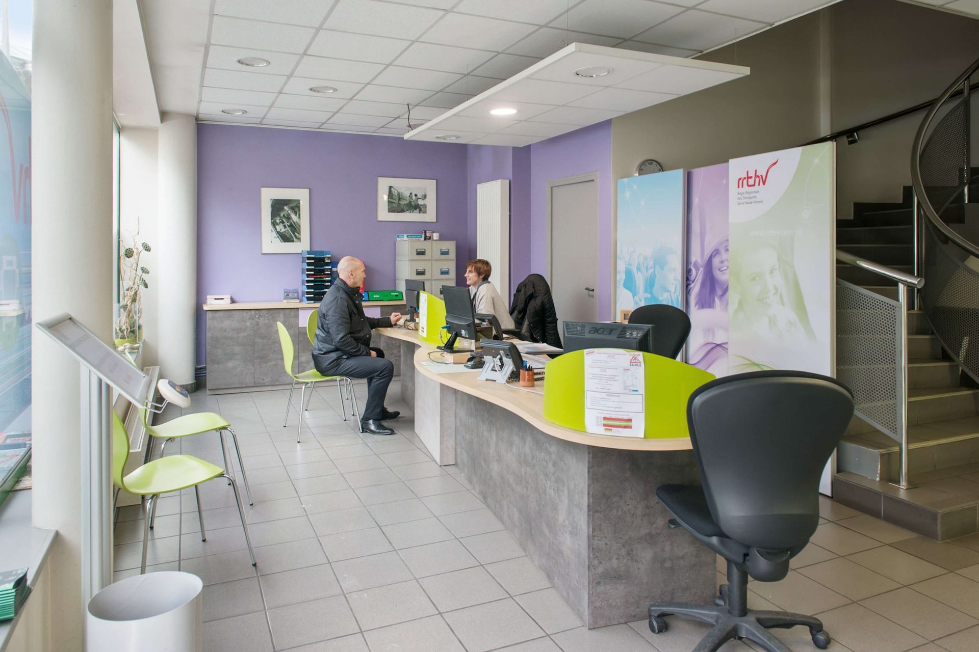 Espace d'accueil : Deux personnes à un bureau, un mur violet et un escalier.