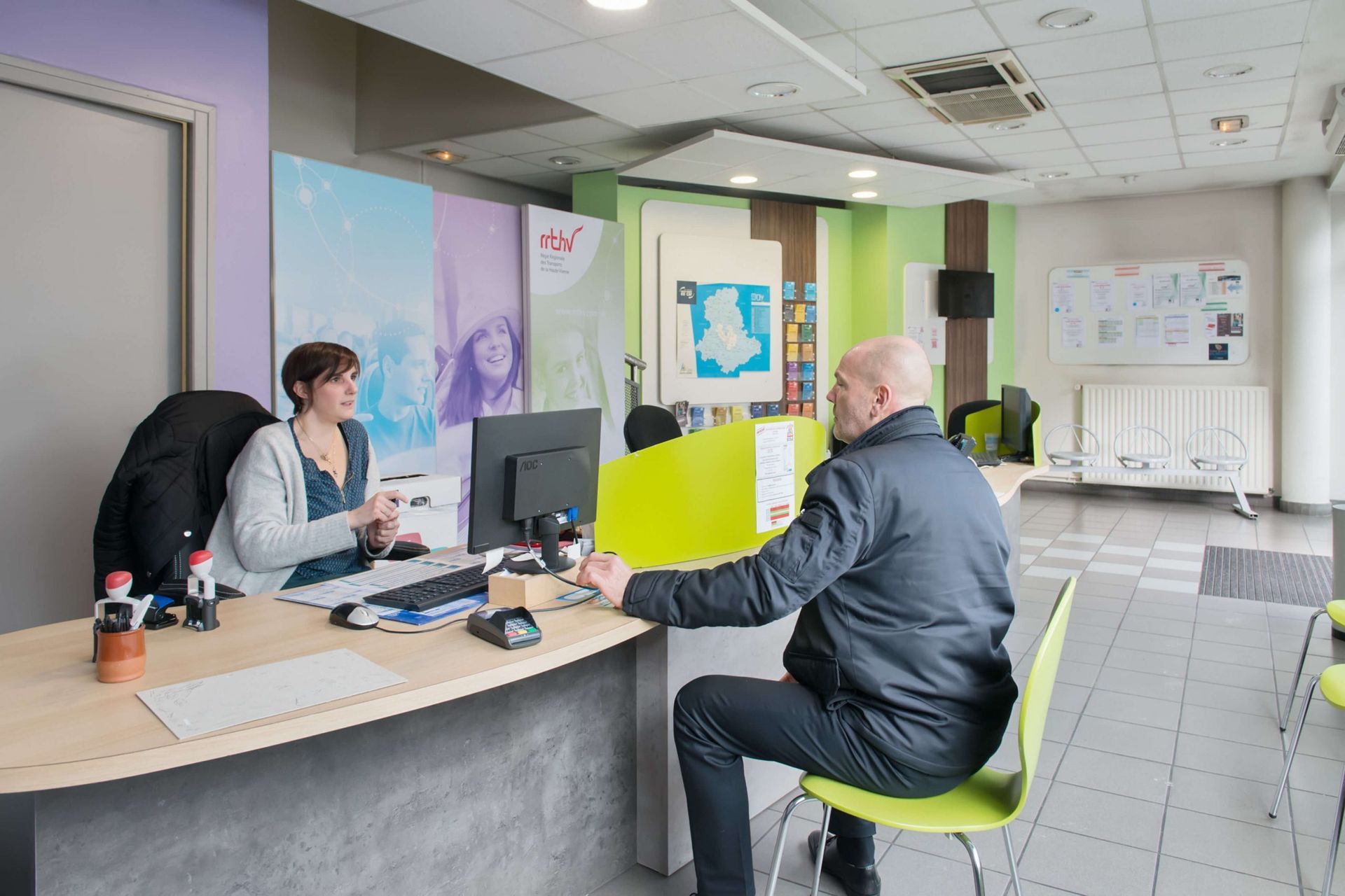 Un homme est assis à un bureau et parle à une personne derrière lui. Ambiance de bureau, tons verts et clairs.