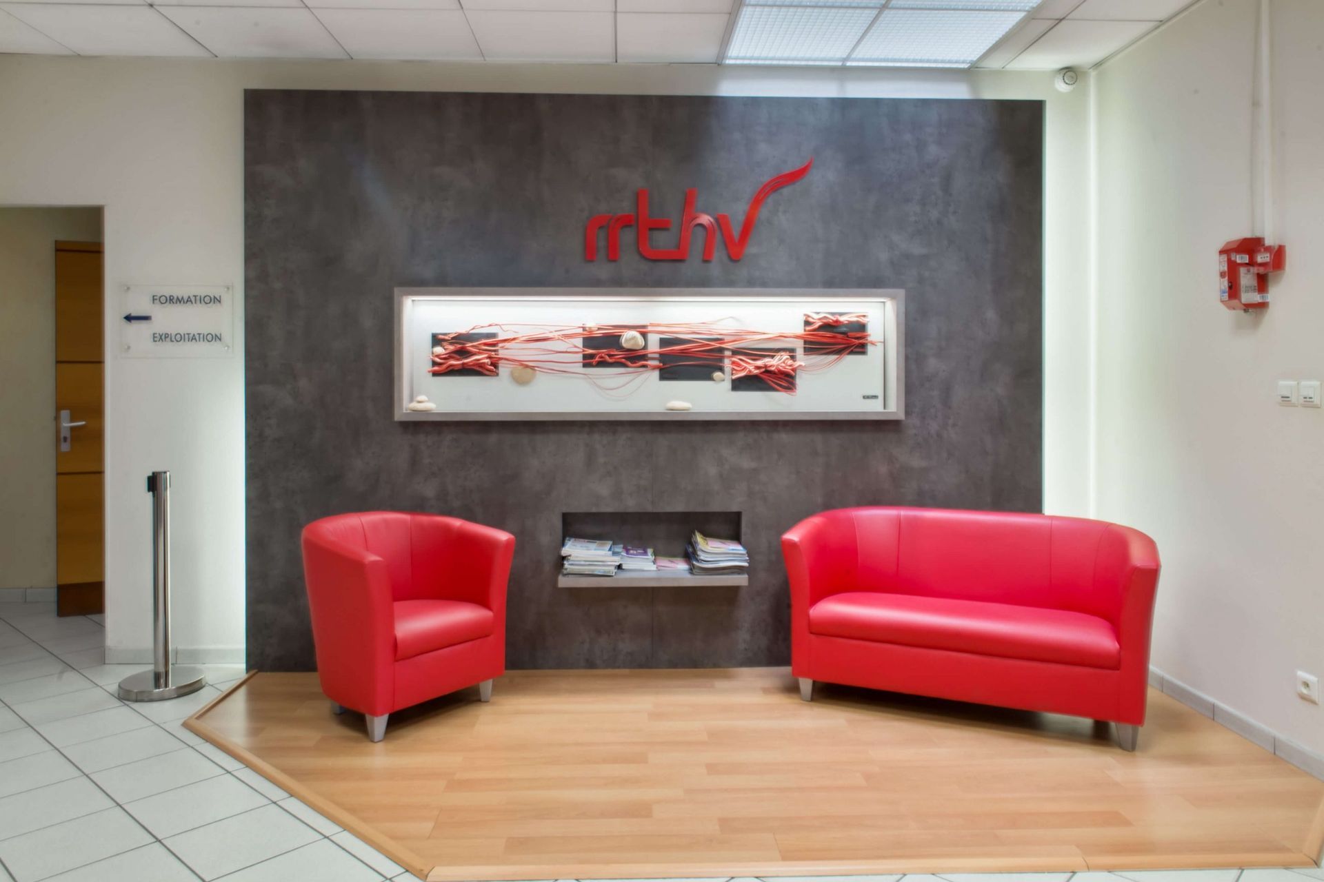 Des sièges rouges dans un espace d'accueil avec un mur d'accent gris foncé, un logo, des photos encadrées et un parquet.