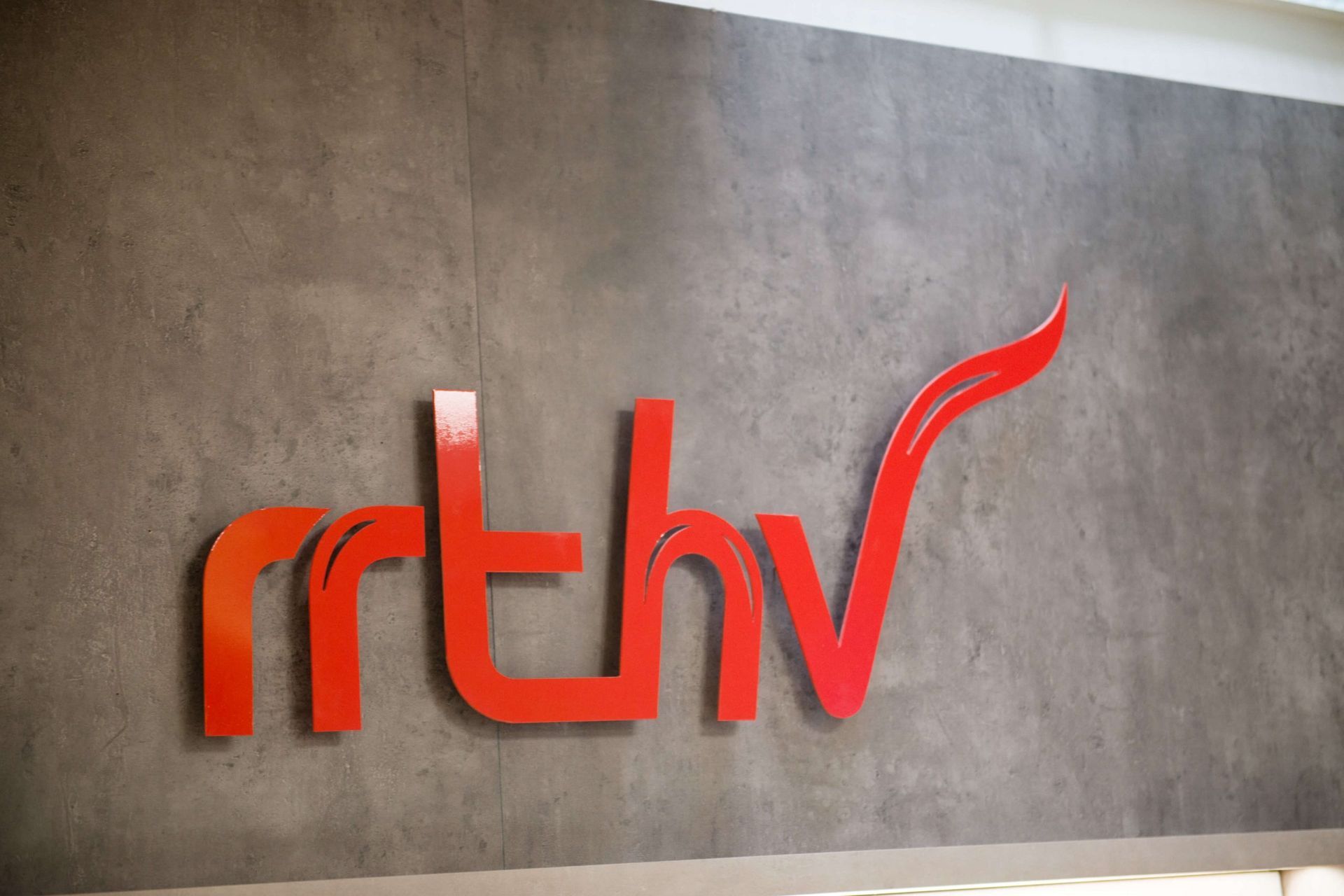 Logo rouge « rthv » sur un mur gris texturé.