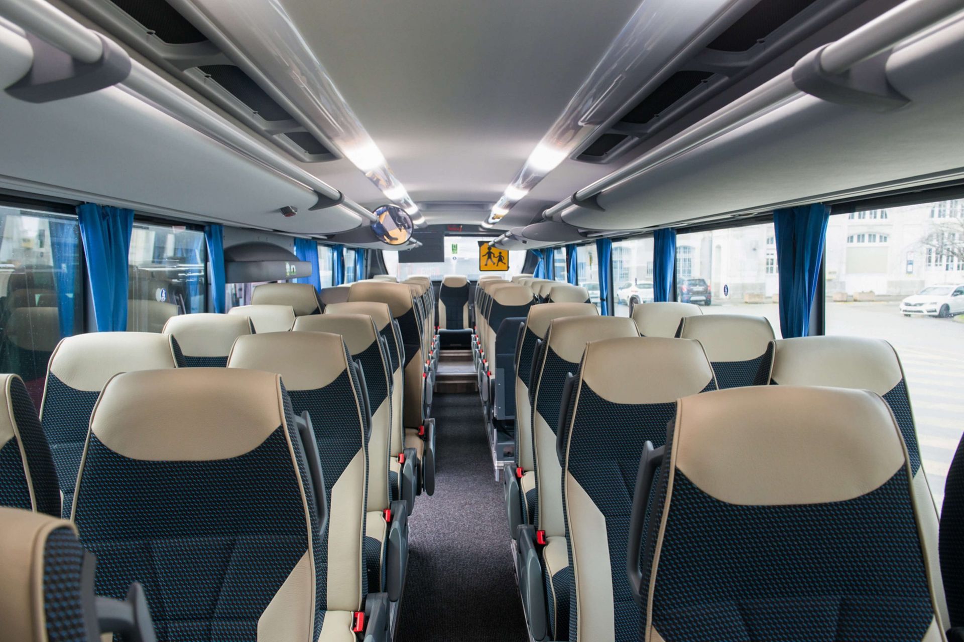 Vue intérieure d'un bus avec des rangées de sièges beiges et bleus, des éclairages au plafond et des rideaux bleus aux fenêtres.