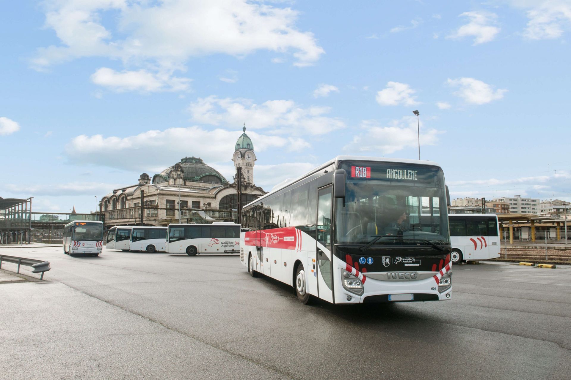 Des bus stationnés à une gare sous un ciel nuageux. Un bâtiment à dôme se profile à l'arrière-plan.