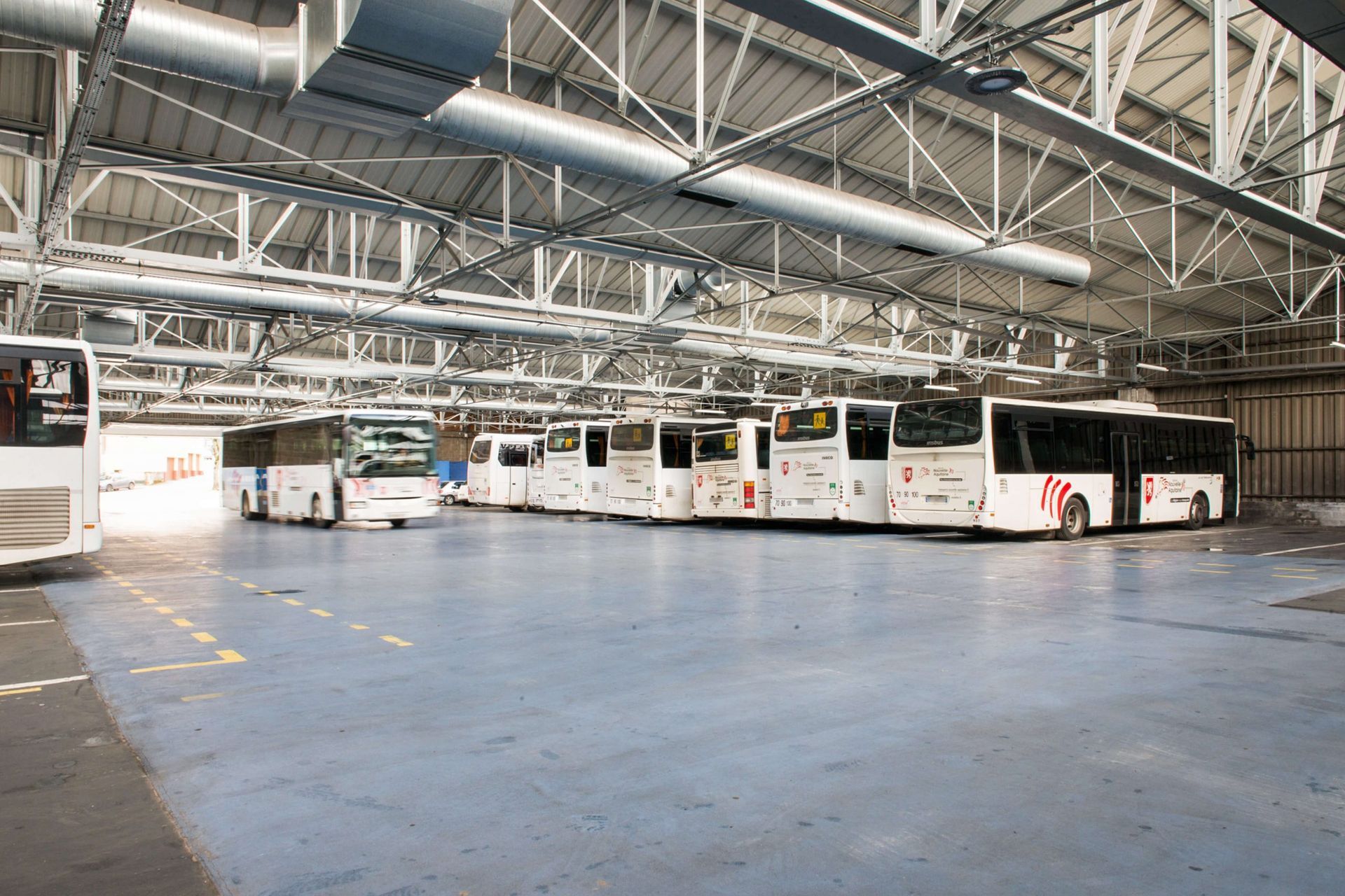 Des bus sont stationnés à l'intérieur d'un grand dépôt au plafond métallique élevé. Le sol est bleu.