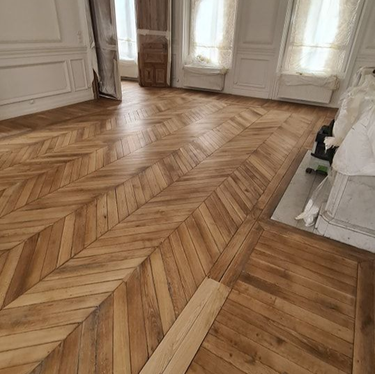 Une pièce vide avec un parquet à chevrons et des murs blancs, éclairée par de grandes fenêtres.