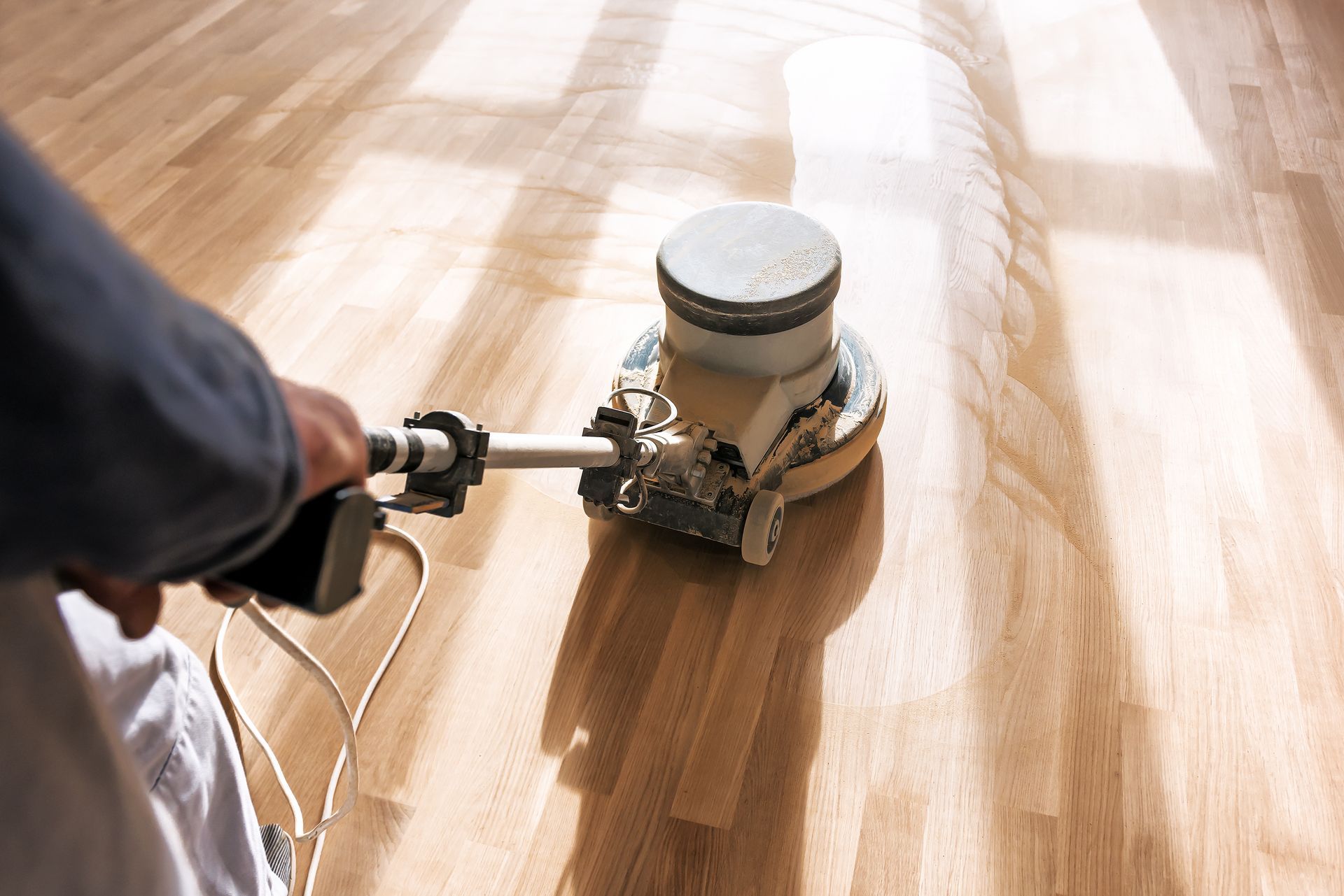 Une personne utilise une polisseuse pour poncer un parquet dans une pièce ensoleillée.