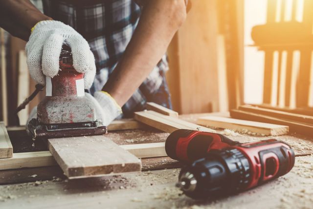 Une personne ponce du bois avec une ponceuse portative dans un atelier, avec des outils et des planches sur la table.