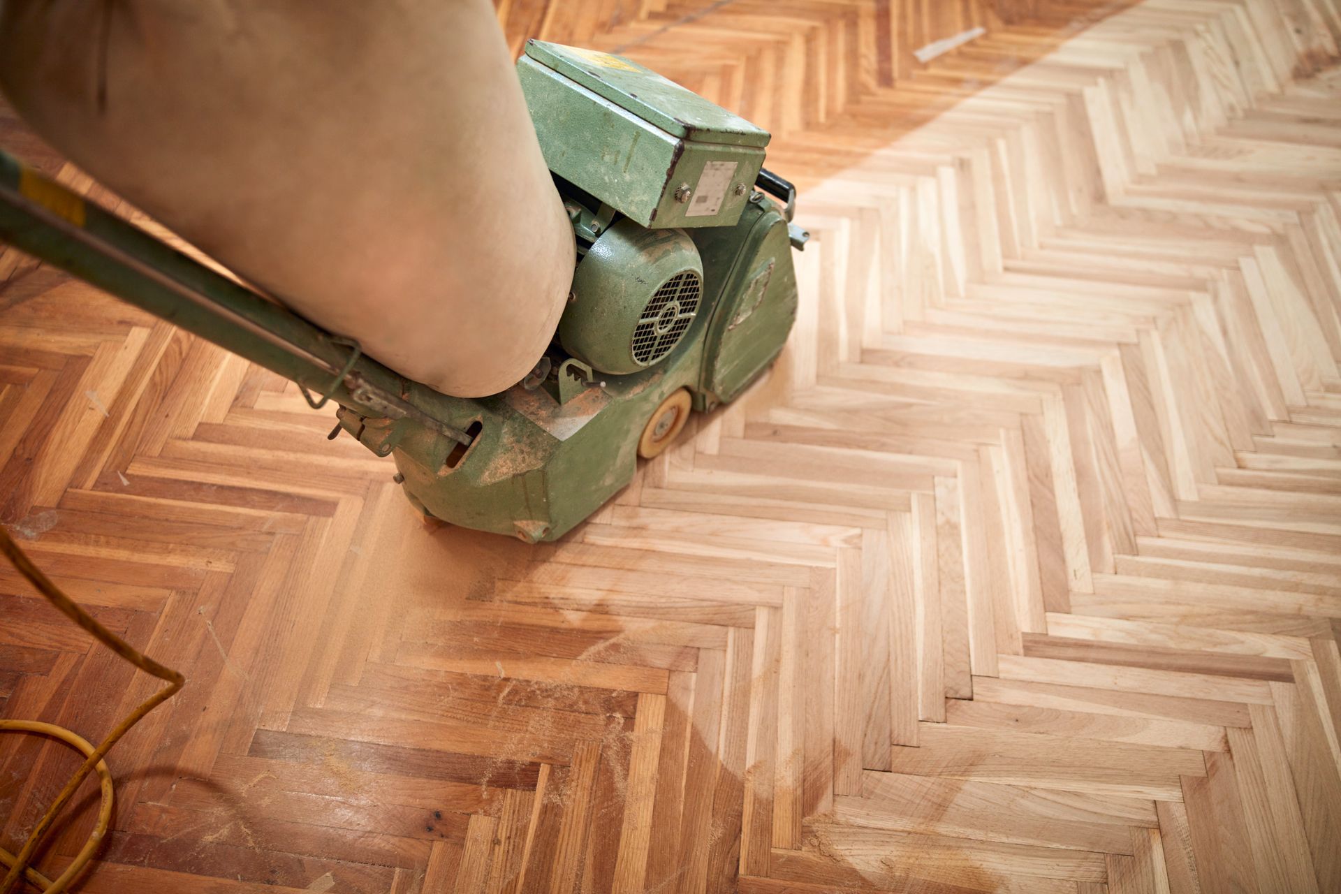 Une personne ponce un parquet à chevrons avec une ponceuse à parquet verte.