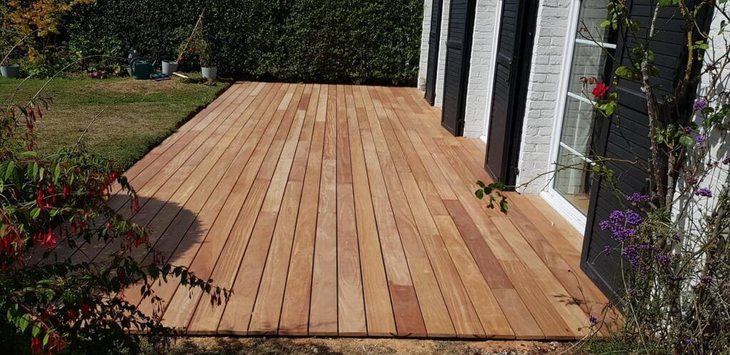 Une terrasse en bois à l'arrière d'une maison, bordée de haies.