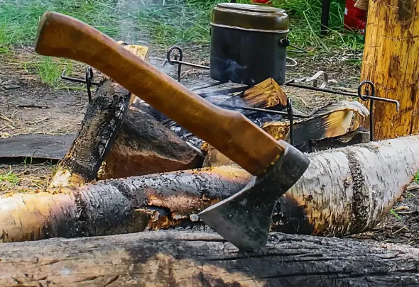 Hacha clavada en un tronco frente a una hoguera.