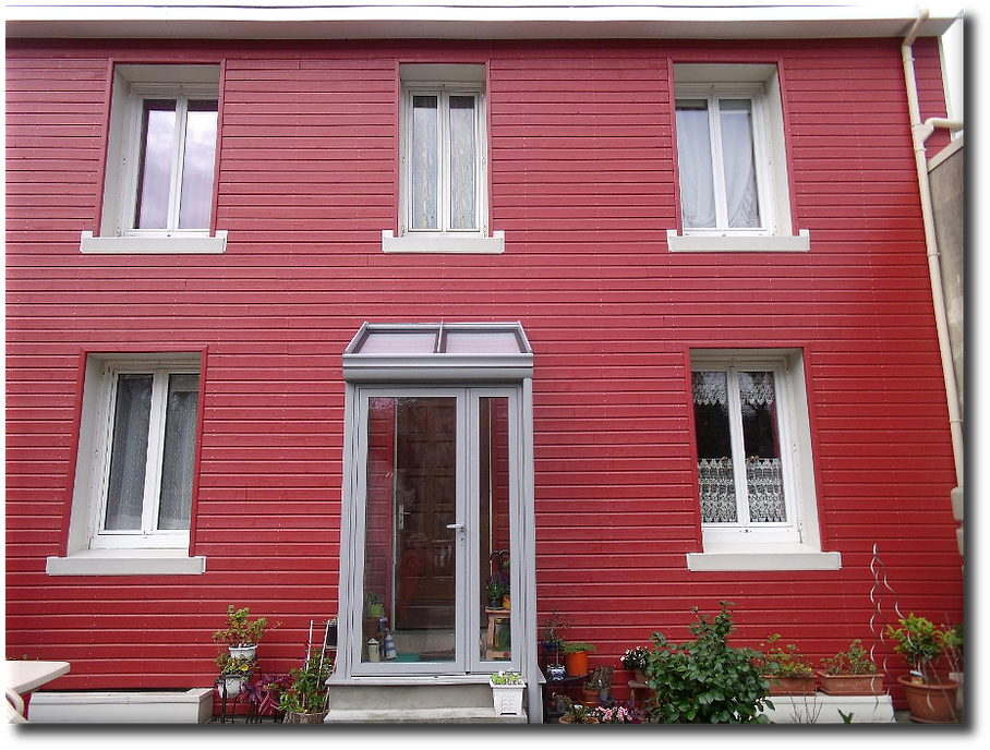 Maison rouge avec six fenêtres, boiseries blanches et entrée vitrée. Plantes en pots en dessous.