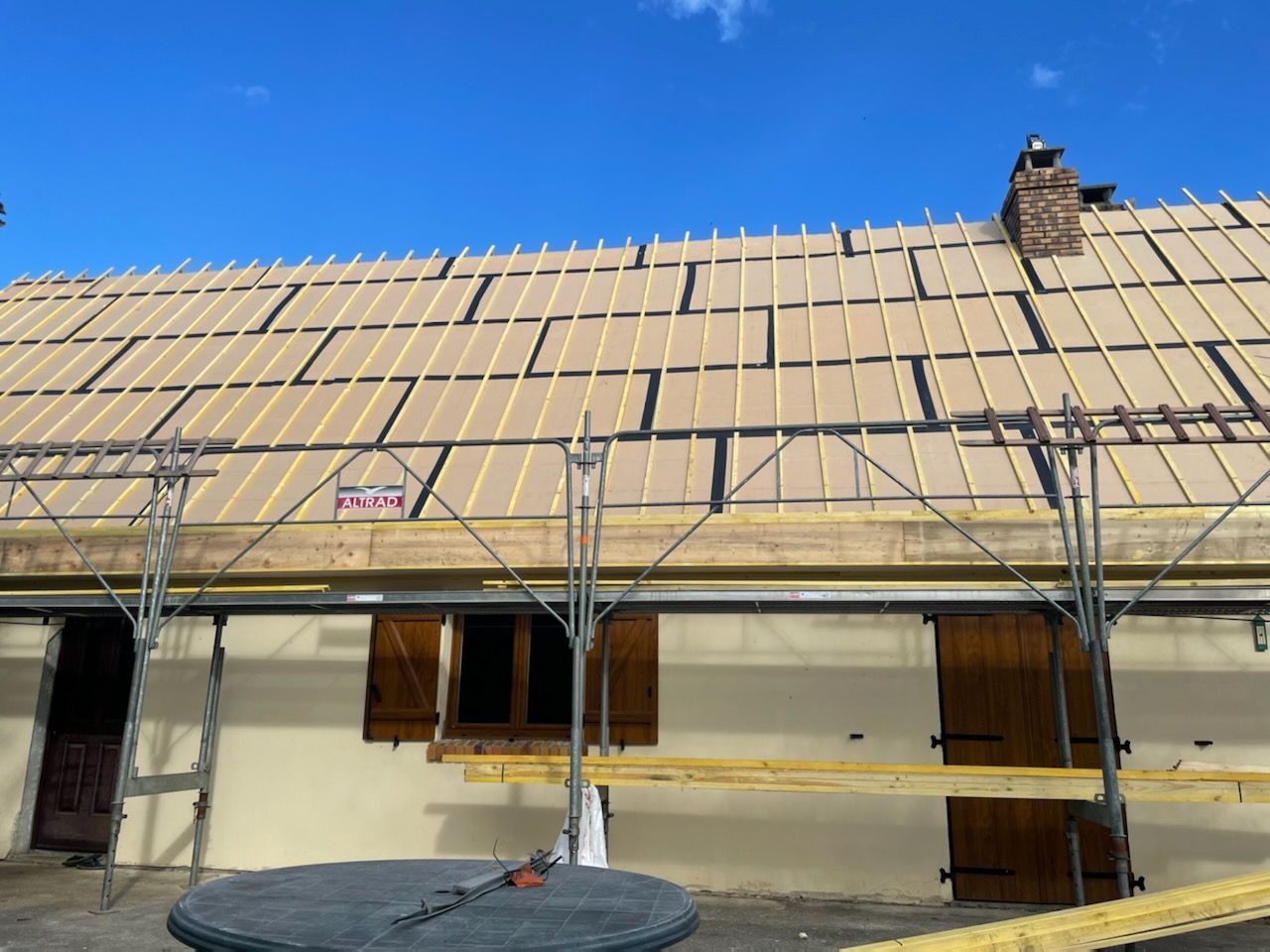 Remplacement de toiture en cours sur une maison avec échafaudage et volets en bois marron sous un ciel bleu.