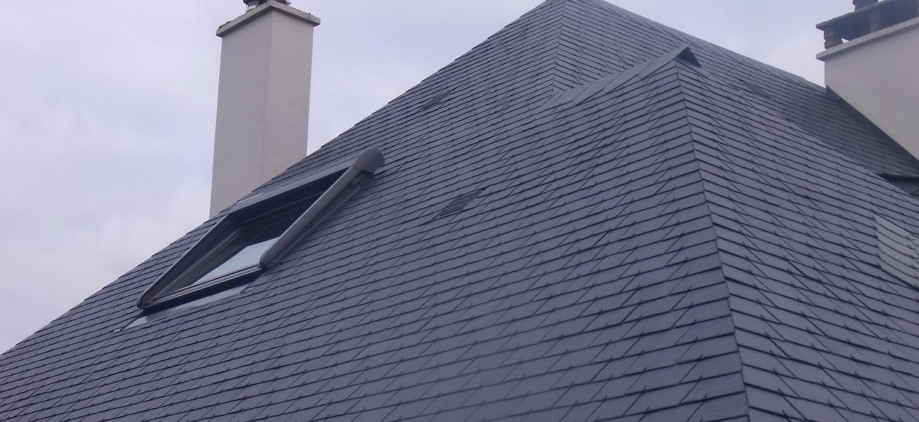Toit en ardoise grise avec puits de lumière et cheminée contre un ciel nuageux.