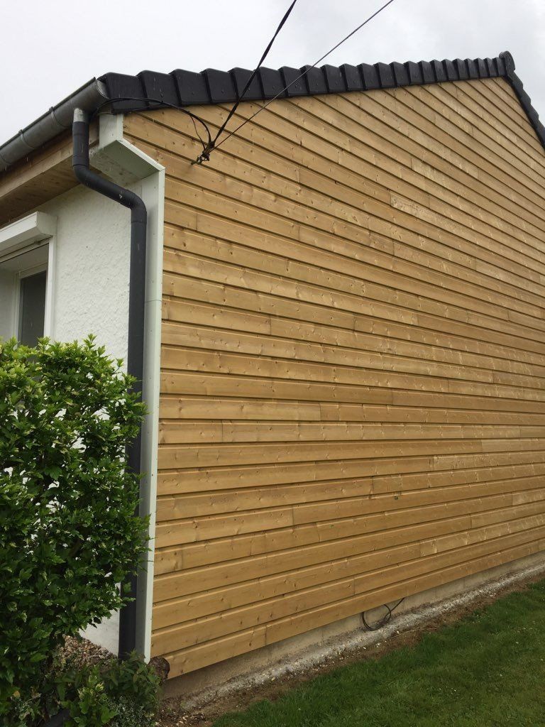 Façade de maison en bois avec moulures, gouttières et antenne noires. Herbe verte et buissons au premier plan.