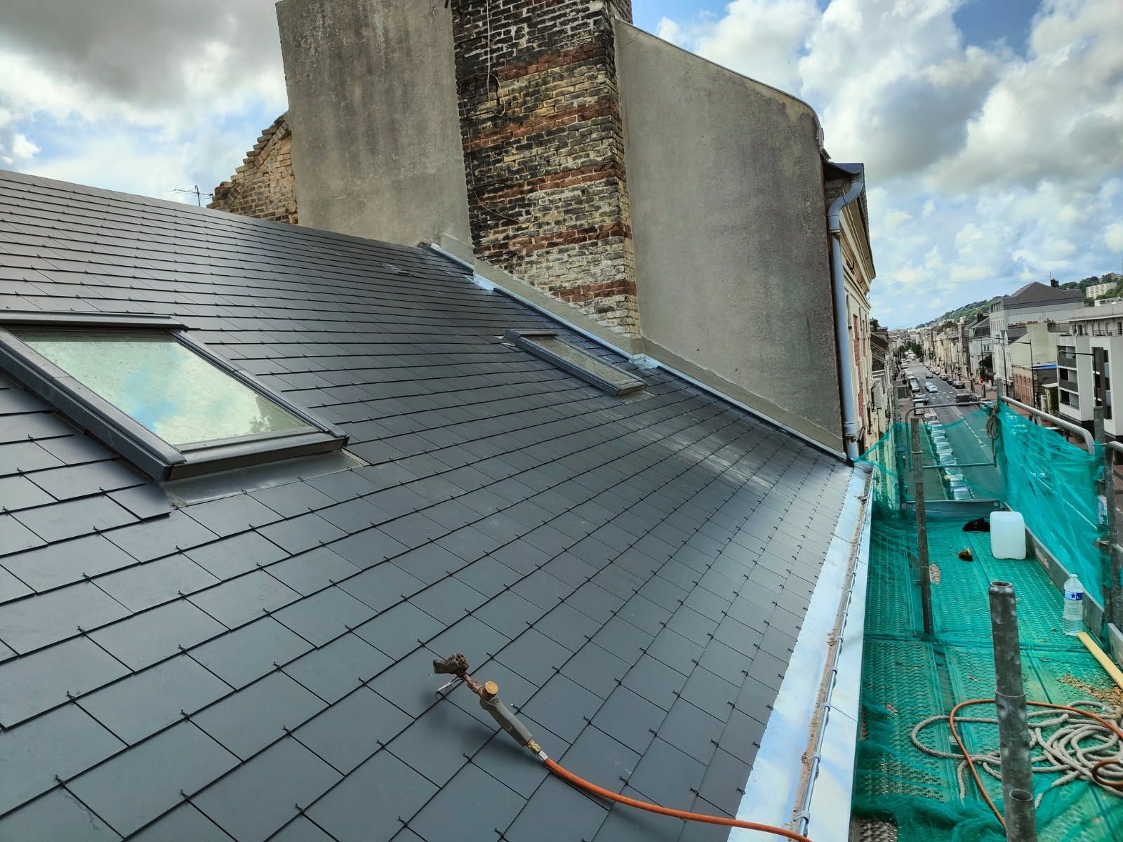 Toiture en ardoise gris foncé avec deux puits de lumière ; échafaudage et rue en arrière-plan.