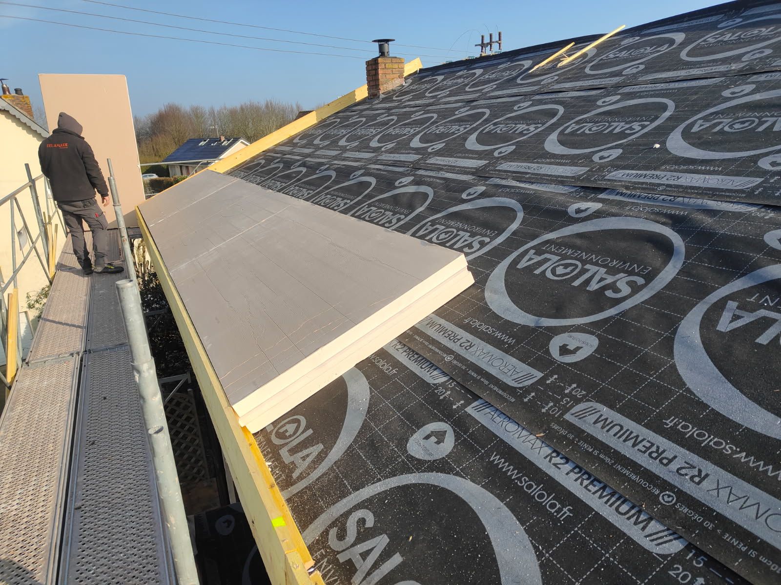 Un couvreur pose de l'isolant sur un toit en pente. Bâche noire et planches visibles.