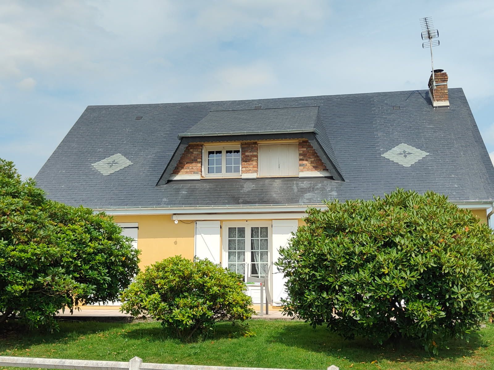 Maison jaune avec toit en ardoise, lucarne et cheminée. Buissons verts devant.