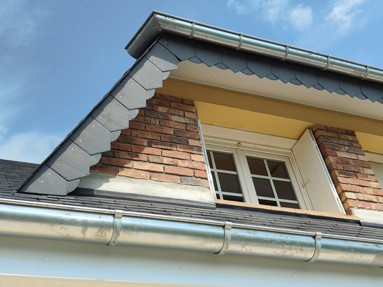 Lucarne avec toit en ardoise foncée, revêtement en brique, cadre blanc et gouttières argentées sur fond de ciel bleu.