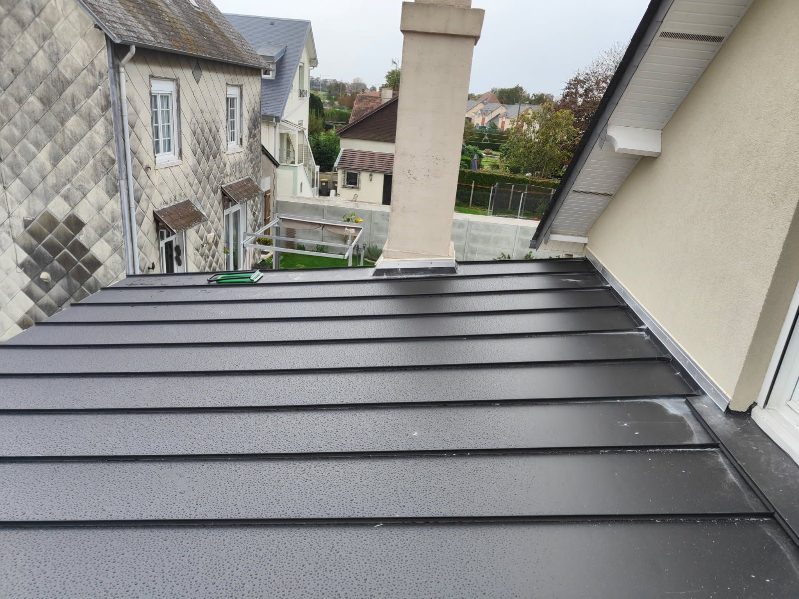 Toiture en métal foncé nouvellement installée avec gouttes d'eau, cheminée et bâtiments voisins.