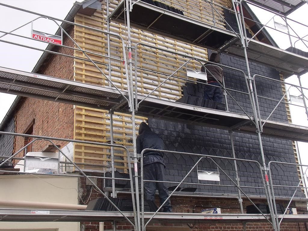 Des ouvriers sur un échafaudage installent un revêtement noir sur un bâtiment en briques.