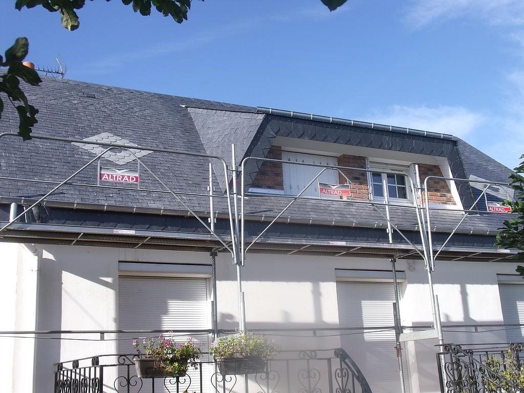 Maison avec toit en ardoise, échafaudage et fenêtres à volets contre un ciel bleu.