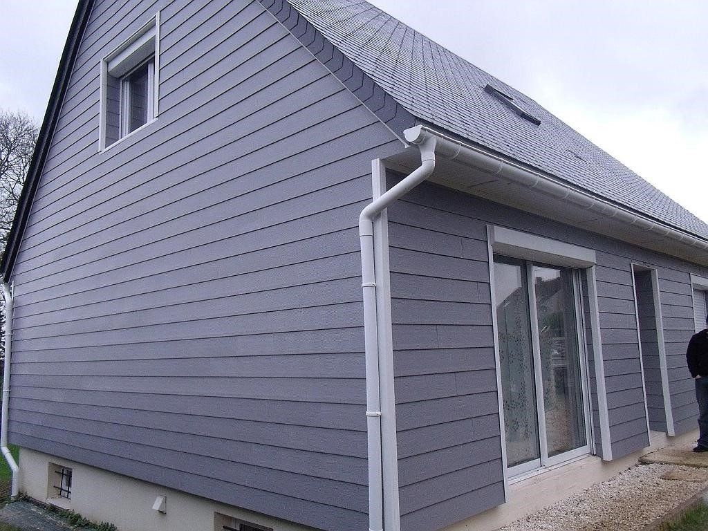 Maison aux façades grises avec moulures et gouttières blanches. Une personne se tient près des fenêtres.